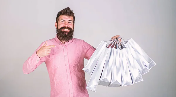 Concepto de recomendación. Hipster en la cara sonriente recomienda comprar. Un tipo de compras en temporada de ventas, apuntando a las bolsas. Hombre con barba y bigote lleva montón de bolsas de compras, fondo gris —  Fotos de Stock