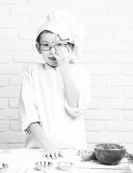 Młody chłopak małych cute kucharz kucharz w biały uniform i kapelusz na twarz Witraż mąki z okularami stojący w pobliżu tabeli z wałkiem czerwony bowl i przytrzymując cookie cutter star na tle ściany Cegła — Zdjęcie stockowe