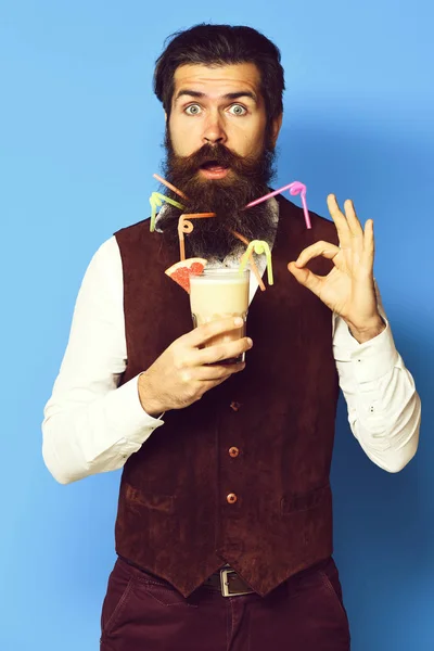 Homem barbudo bonito com barba longa e bigode tem cabelo elegante no rosto surpreso segurando vidro de coquetel alcoólico em jaqueta de couro de camurça vintage no fundo do estúdio azul — Fotografia de Stock