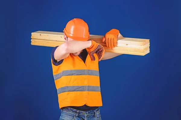 Carpenter, woodworker, labourer, builder on tired face carries wooden beam on shoulder. Tired labourer concept. Man in helmet and protective gloves wiping sweat from forehead, blue background — Stock Photo, Image