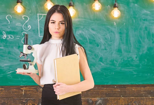 Profesor de biología tiene libro y microscopio. Dama en ropa formal con la cara tranquila en el aula. Concepto de biología. Lady científica sostiene libro y microscopio, pizarra en el fondo, espacio de copia — Foto de Stock