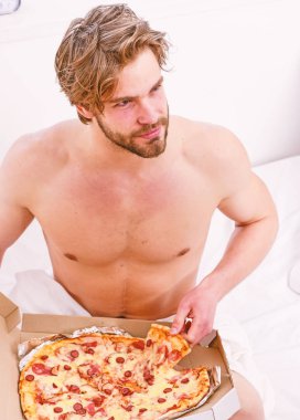 Bir yatağa evde döşeme sırasında tembel kaslı adam yeme pizza portresi. Gömleksiz seksi adam yatakta pizza ile görüntü kırpılmış. Ara diyet kavramı.