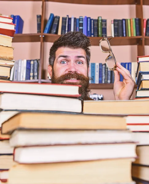 Enseignant ou élève barbu porte des lunettes, s'assoit à table avec des livres, déconcentré. Homme au visage surpris entre des piles de livres à la bibliothèque, étagères sur fond. Concept de recherche scientifique — Photo