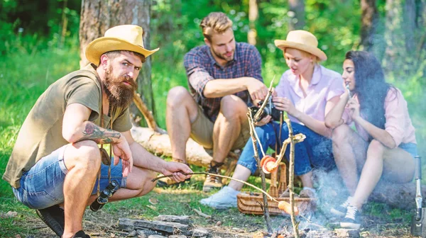 Hipster roasting sausage while friends speaking sharing impression and watching photos on camera. Friends group tourist relaxing near bonfire. Tourists having snack time with roasted over fire food — Stock Photo, Image
