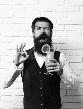 Uzun sakallı ve bıyıklı yakışıklı adamın komik yüzünde şık saçları var. Elinde beyaz tuğla duvar arka planında klasik süet deri yelek ve alkollü içecek bardağı var.