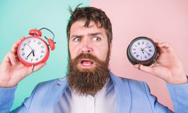Changing time zones affect health. Time zone. Does changing clock mess with your health. Man bearded hipster hold two different clocks. Guy unshaven puzzled face having problems with changing time clipart