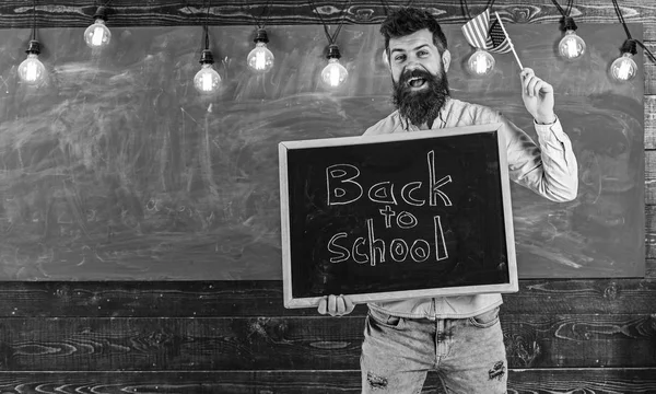 Lehrerin hält Tafel mit Schriftzug zurück zur Schule und Fahne der USA. Amerikanisches Schulkonzept. Mann mit Bart auf lächelndem Gesicht begrüßt amerikanische Schule, Kreidetafel im Hintergrund — Stockfoto