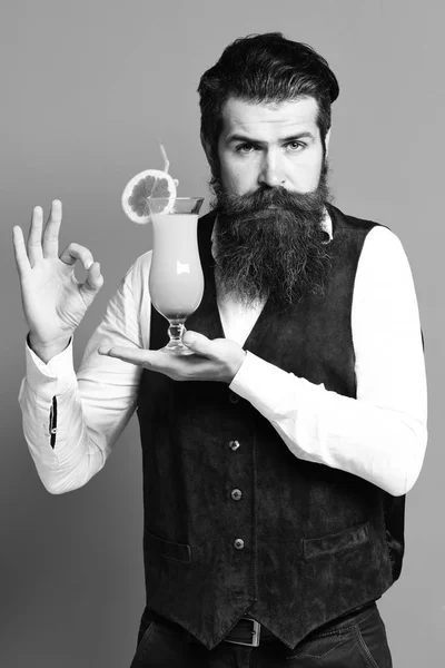 handsome bearded man with long beard and mustache has stylish hair on serious face holding glass of alcoholic cocktail in vintage suede leather waistcoat on red studio backgroun