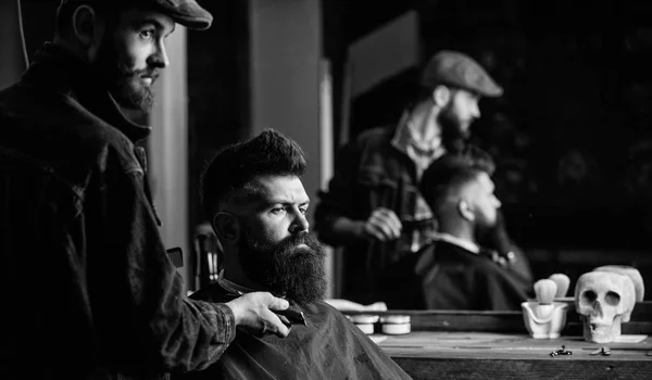 Hipster with beard covered with cape serving by professional barber in stylish barbershop. Grooming concept. Barber busy with grooming beard of hipster client, mirror reflexion on background — Stock Photo, Image