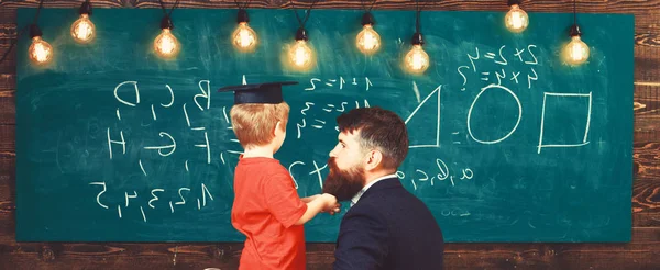 老师带着胡子, 父亲在教室里教小儿子, 黑板上的背景。男孩, 孩子在研究生盖帽听老师, 黑板在背景, 后方看法。学校概念的准备 — 图库照片