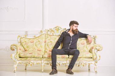 Sakallı ve bıyıklı lüks oturma odasında boş zaman harcıyor. Hipster düşünceli yüzünde yalnız oturuyor. Zengin ve yalnız kavramı. Lüks daire sahibi beyaz duvar arka koltukta oturur