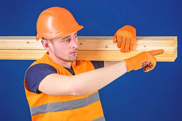 Tischler, Holzarbeiter, kräftiger Bauarbeiter auf dem Gesicht trägt Holzbalken auf der Schulter. Mann mit Helm, Schirmmütze und Schutzhandschuhen, die Richtung weisen, blauer Hintergrund. Holzwerkstoffkonzept — Stockfoto