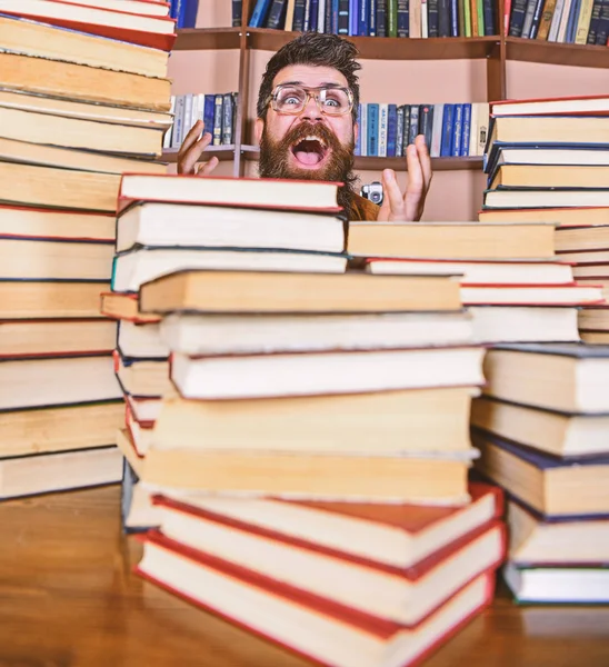 Вчитель або учень з бородою носить окуляри, сидить за столом з книгами, дефокусовані. Наукова концепція відкриття. Чоловік на збудженому обличчі між купами книг у бібліотеці, книжкові полиці на фоні — стокове фото
