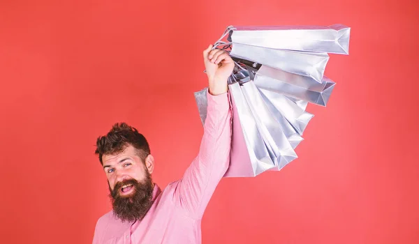 Hombre con barba y bigote sostiene bolsas de compras, fondo rojo. Hombre de compras en temporada de ventas con descuentos. Hombre hipster caucásico sosteniendo montón de bolsas de compras. Concepto de compras —  Fotos de Stock