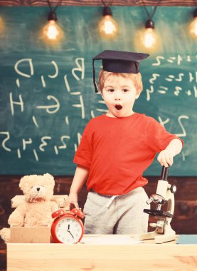 Çocuk, öğrenci mikroskop yakınındaki şok yüzünde. Sınıf mikroskobu, kara tahta yakınındaki yüksek lisans şapkalı çocuk arka plan üzerinde evlat. İlk eski okumak, eğitim isteyen. Harika çocuk kavramı