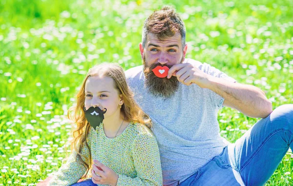 Gender roles concept. Family spend leisure outdoors. Dad and daughter sits on grassplot, grass on background. Father posing with lips and child posing with beard photo booth attribute — Stock Photo, Image