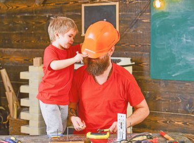 Baba, ana okul atölyesinde farklı araçları kullanmak için kask öğretim oğlu sakallı. Evlat, çocuk neşeli cıvata veya vida, baba ile handcrafting sırasında eğlenmek tutar. Tamirci kavramı