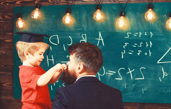 Individuell utbildning koncept. Liten pojke leker med skägg av mannen bredvid honom. Vuxna vände tillbaka och ler barnet i examen cap sidovy. Ung kille och liten unge framför gröna styrelsen — Stockfoto