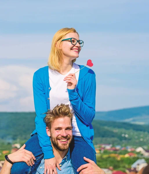 Homme porte petite amie sur les épaules, fond de ciel. Concept romantique de rendez-vous. Femme tient le cœur sur bâton symbole d'amour. Couple amoureux marche journée ensoleillée en plein air. Couple heureux date avoir du plaisir ensemble — Photo