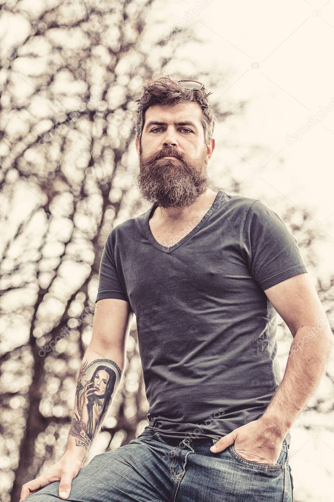 Tired middle-aged man with long beard standing in park on warm spring day. Muscular brutal man with tattooed arm wearing dark blue T-shirt and denim jeans walking in urban scene