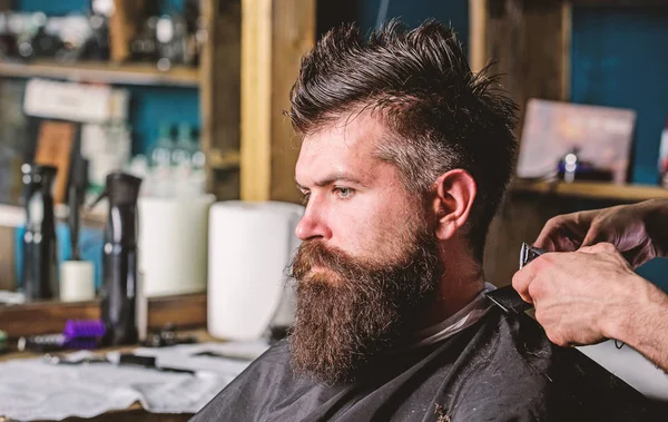 Hands of barber with clipper and comb, close up. Hipster bearded client getting hairstyle. Barbershop concept. Man with beard in hairdressers chair, salon background. Barber works with hair clipper