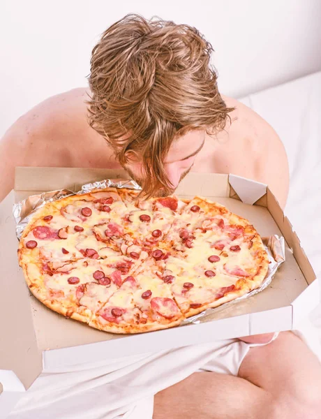 Man bearded handsome guy eating cheesy food for breakfast in bed. Shirtless handsome young man with pizza on bed. Student is at home on the bed in a bright apartment eating a tasty pizza.