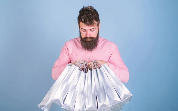 Man with trendy hairstyle and beard holding silver bags. Hipster in pink shirt with curious look isolated on blue background. Concentrated bearded man opening presents, birthday celebration concept — Stock Photo, Image