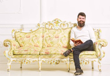 Sakal ve bıyık oturur barok tarzı kanepe, tutar kitap, beyaz duvar arka plan üzerinde olan adam. Adam zevk ile eski kitap okuma. Okuyucu kavramı. Maço yüz kitap okuma konusunda tutkulu gülümseyen üzerinde