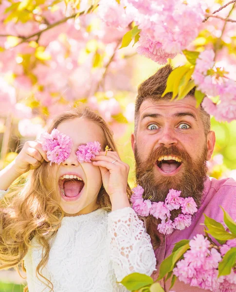 Father and daughter on happy face play with flowers as glasses, sakura background. Girl with dad near sakura flowers on spring day. Child and man with tender pink flowers in beard. Family time concep — Stock Photo, Image