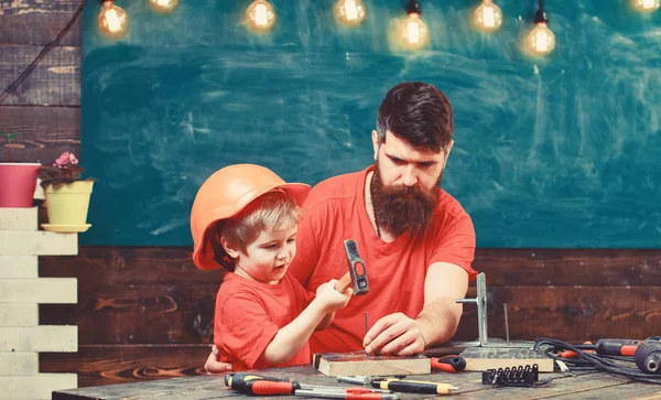 Ребенок, занятый в защитном шлеме, учится пользоваться молотком вместе с отцом. Отец с бородой учит маленького сына пользоваться инструментами, молотком, доской на заднем плане. Маленький ассистент — стоковое фото