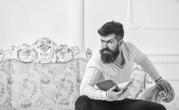 Homme avec barbe et moustache assis sur un canapé de style baroque, tient livre, fond mural blanc. Macho sur le visage strict fini livre de lecture. Un type qui pense à la littérature. Réflexions sur le concept littéraire — Photo