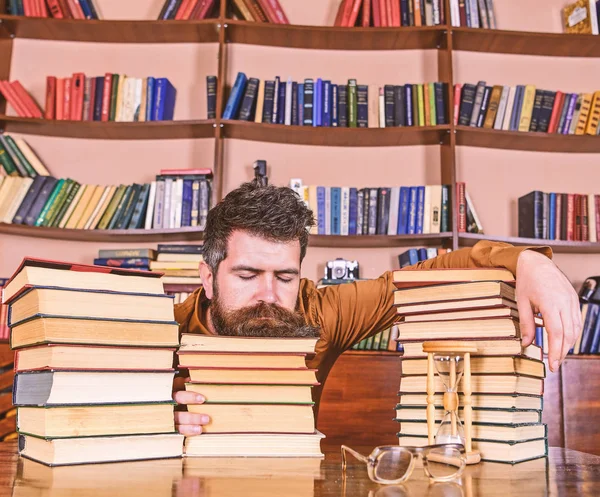 Homem no rosto dormindo estava entre pilhas de livros, adormecer enquanto estudava na biblioteca, estantes de livros no fundo. Conceito demasiado estudado. Professor ou estudante com barba adormecer em livros, desfocado — Fotografia de Stock