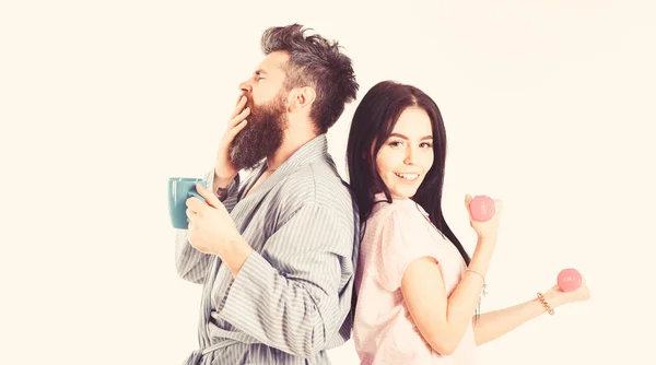 Couple, family on sleepy faces, full of energy. Couple in love in pajama, bathrobe stand isolated on white background. Girl with dumbbell, man with coffee cup. Alternative lifestyle concept