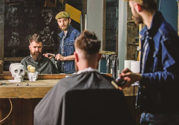 Barber preparing hair clipper for bearded man, barbershop background. Hipster lifestyle concept. Hipster client covered with cape getting haircut. Barber with clipper and brutal bearded client