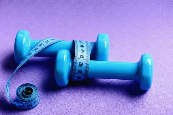 Dumbbells and measure tape in cyan color on purple background.
