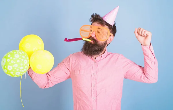 Cheerful bearded man in birthday cap blowing party whistle, fun time, happiness concept. Artist in big crazy glasses holding colorful balloons. Man with hipster beard and mustache celebrating