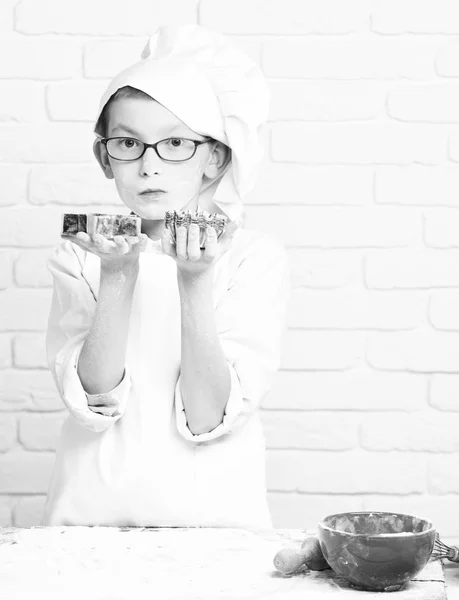 Young boy small cute cook chef in white uniform and hat on stained face flour with glasses standing near table with rolling pin red bowl and holding cookie cutter stars on brick wall background — Stock Photo, Image