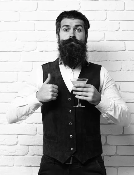 Hombre barbudo guapo con barba larga y bigote tiene el pelo elegante en la cara divertida celebración de vidrio de bebida alcohólica en chaleco de cuero de gamuza vintage en fondo de estudio de pared de ladrillo blanco — Foto de Stock