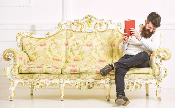 남자는 즐거움 함께 오래 된 책을 읽고입니다. 유머 문학 개념입니다. 바로크 스타일 소파, 보유도 서, 흰 벽 배경에 수염과 콧수염 앉아와 남자. 얼굴 책을 읽고 웃 고에 사나이 — 스톡 사진