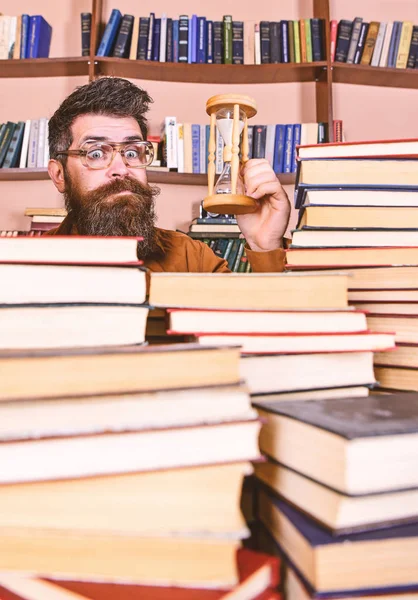 Homme au visage surpris tient sablier tout en étudiant, étagères sur fond. Concept de flux temporel. Enseignant ou étudiant barbu étudiant à la bibliothèque. Homme, scientifique dans les lunettes regarde sablier — Photo