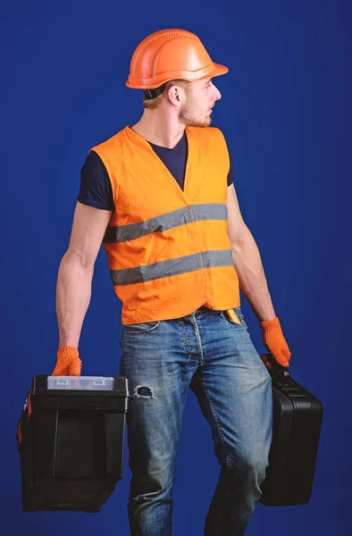 Чоловік в шоломі, жорсткий капелюх тримає коробку інструментів і валізу з інструментами, синій фон. Обладнана концепція ремонтників. Працівник, майстер, ремонтник, будівельник на спокійному обличчі несе сумки з професійними інструментами — стокове фото