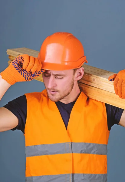 Carpenter, woodworker, strong builder on busy face carries wooden beam on shoulder. Safety and protection concept. Man in protective gloves holds visor of protective hard hat, grey background — Stock Photo, Image