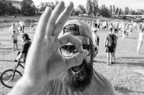 Man cheerful face looks through ok gesture. Man bearded in front of crowd riverside background. Book ticket summer festival in advance. Be sure in your summer vacation. Hipster visiting fest festival