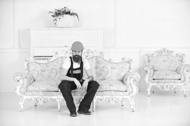 Yükleyici dinlenme kanepeye otur. Break ve dinlenme kavramı. Sakallı adam, Tulum ve kask oturan işçi yorgun, beyaz arka plan kanepe. Kurye mobilya, tehcir hareket ederken rahatlatıcı