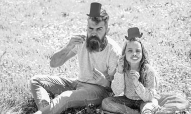 Aristocrates kavramı. Baba ve kızı, grassplot, arka planı yeşil çimenlerin üzerinde oturur. Çocuk ve babası silindir şapka, dudaklar ve boru photo booth öznitelikleri ile poz. Aile harcamak açık havada eğlence
