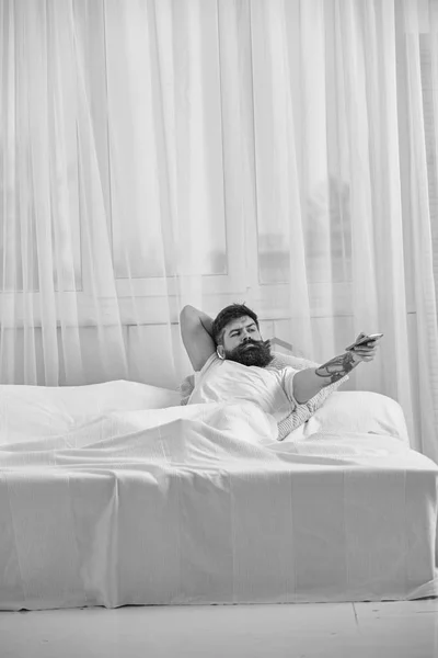 Hombre de camisa acostado en la cama, viendo la televisión, cortinas blancas en el fondo. Macho con barba sostiene el controlador en la mano. Concepto de control remoto. Tipo en cara seria usando control remoto para canal de conmutación —  Fotos de Stock