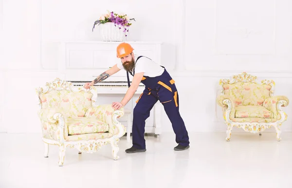 Levering dienstverleningsconcept. Koerier levert meubels in geval van verhuizing uit, verplaatsing. Lader draagt fauteuil. Man met baard, worker in overalls en helm duwt leunstoel, witte achtergrond — Stockfoto