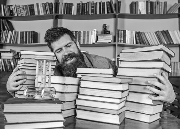 Concepto bibliotecario. Hombre en la cara feliz entre montones de libros, mientras estudia en la biblioteca, estanterías en el fondo. Profesor o estudiante con barba se sienta a la mesa con libros, desenfocado —  Fotos de Stock