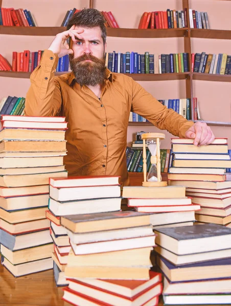 Könyvtáros koncepció. Átgondolt arcán ember között halom könyv, könyvtár, tanulmányai könyvespolcok a háttérben áll. Tanár vagy diák szakáll áll asztalhoz a könyvek, defocused — Stock Fotó