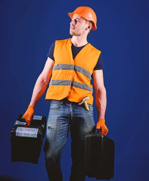 Homem de capacete, chapéu duro segura caixa de ferramentas e mala com ferramentas, fundo azul. Handyman, reparador no rosto sonhador vai e carrega sacos com equipamento profissional. Conceito de reparador profissional — Fotografia de Stock